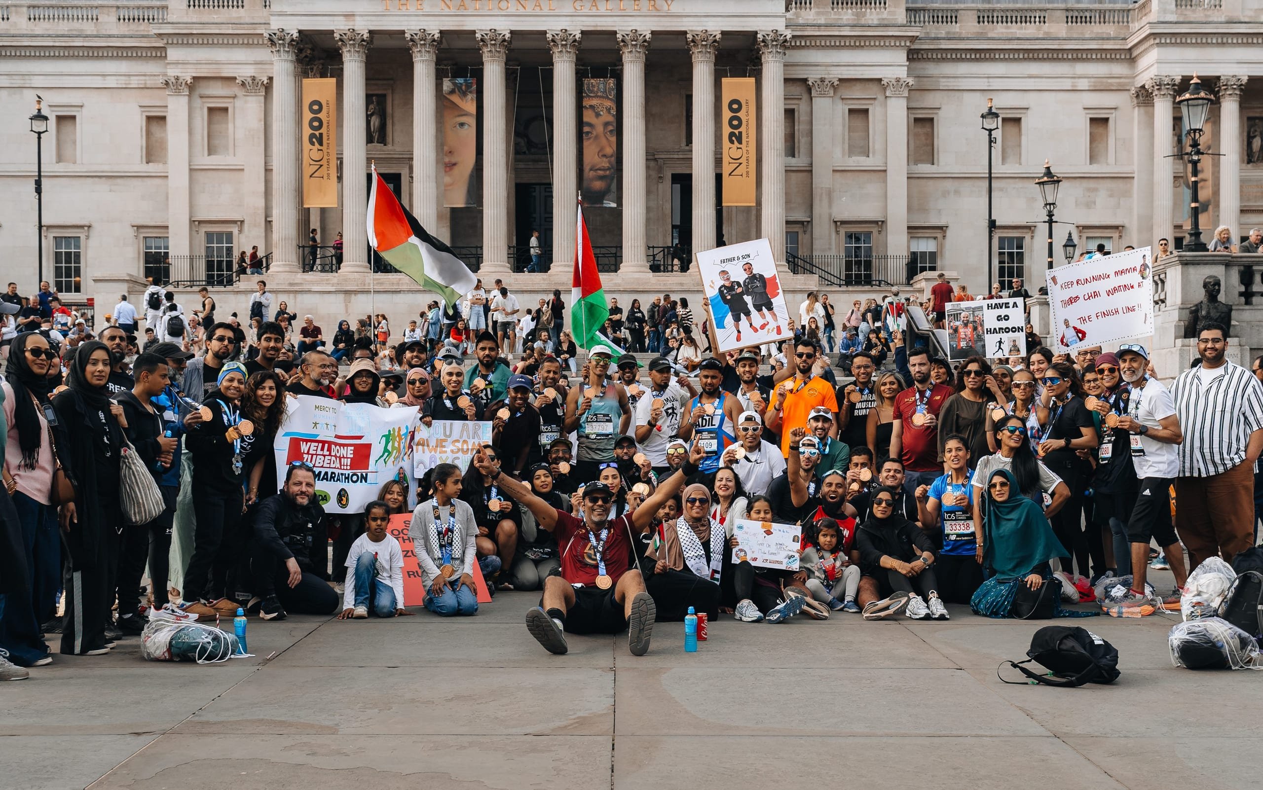 Muslim Runners community — over 50 people gathered at Trafalgar Square after the London Marathon with medals, signs reading 'Have a go Haroon' and 'Keep running', Palestinian flags, and pure collective joy